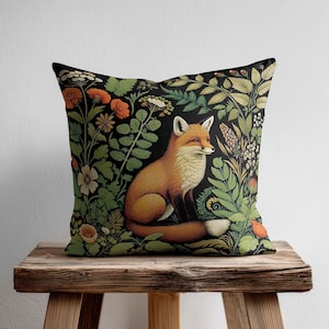 Op de afbeelding: Een vierkant sierkussen met een gedetailleerde illustratie van een rode vos die midden in een weelderige tuin van groen gebladerte, oranje bloemen en witte bloesems op een zwarte achtergrond zit. De vos heeft een witte borst en staartpunt.
