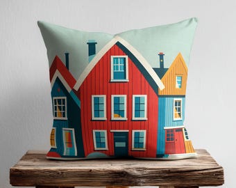 May include: A square throw cushion featuring a colourful illustration of three houses. The central house is red, flanked by a blue house on the left and a yellow house on the right. Each house has multiple rectangular windows and a dark blue door.