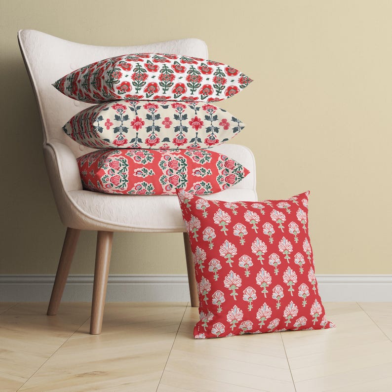 Puede incluir: Cuatro cojines decorativos con patrones florales en tonos rojos, blancos y verdes. Los cojines est&aacute;n apilados en un sill&oacute;n blanco con patas de madera.