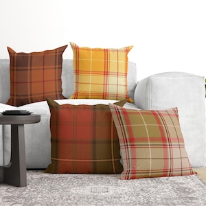 May include: Four square throw pillows featuring various plaid patterns in warm autumn tones. One pillow is dark brown, one is bright yellow, one is deep red and brown, and one is a mix of tan, red, and olive green. Each pillow has a distinct tartan design.