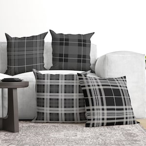 May include: Four square throw pillows featuring various shades of gray, black, and white in distinct plaid and tartan patterns. Each pillow displays a unique grid design with intersecting lines of varying thickness, creating a cohesive monochromatic set.