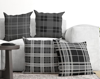May include: Four square throw pillows featuring various shades of gray, black, and white in distinct plaid and tartan patterns. Each pillow displays a unique grid design with intersecting lines of varying thickness, creating a cohesive monochromatic set.