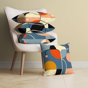 May include: A stack of four decorative throw pillows with abstract geometric patterns in shades of blue, yellow, orange, pink, and black. The pillows are arranged on a white armchair.