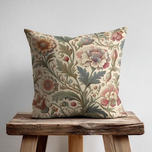 May include: A square throw pillow featuring a cream-colored background with a dense, intricate botanical pattern. The design displays large, multi-petaled flowers in shades of muted pink, peach, and orange, intertwined with detailed green foliage.