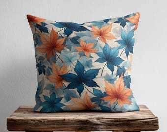 May include: A square throw cushion featuring a repeating pattern of maple leaves in shades of deep blue, light blue, and warm orange against a cream-coloured background. The leaves are arranged in an overlapping design across the entire surface.
