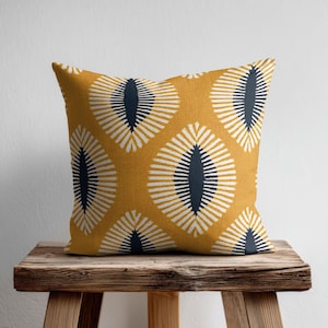 May include: A square throw pillow featuring a mustard yellow background with a repeating pattern of dark navy blue almond-shaped centers surrounded by radiating white lines that create a bold, geometric, and symmetrical visual effect.