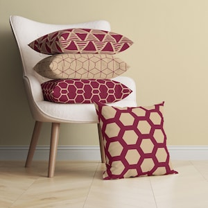 Könnte beinhalten: Vier dekorative Kissen mit geometrischen Mustern in den Farben Burgunderrot und Beige. Die Kissen sind auf einem weißen Sessel mit Holzbeinen gestapelt.