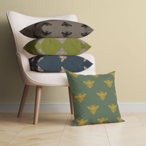 Puede incluir: Una colecci&oacute;n de cojines decorativos con dise&ntilde;os de abejas. Los cojines son de varios colores, incluyendo verde oliva, azul marino y topo. Los dise&ntilde;os de abejas son en colores contrastantes, como dorado y negro. Los cojines est&aacute;n colocados sobre una silla blanca.