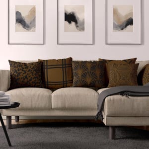 Könnte beinhalten: Ein beigefarbenes Sofa mit fünf Kissen. Die Kissen haben verschiedene geometrische Muster in Schwarz und Braun. Das Sofa steht vor einer weißen Wand mit drei gerahmten abstrakten Gemälden.