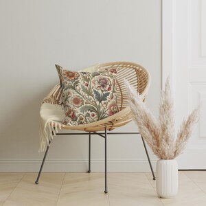 May include: A square throw pillow featuring a detailed botanical pattern with muted red, orange, and green floral motifs on a cream background, resting on a round rattan chair with thin black metal legs and a draped off-white throw blanket.