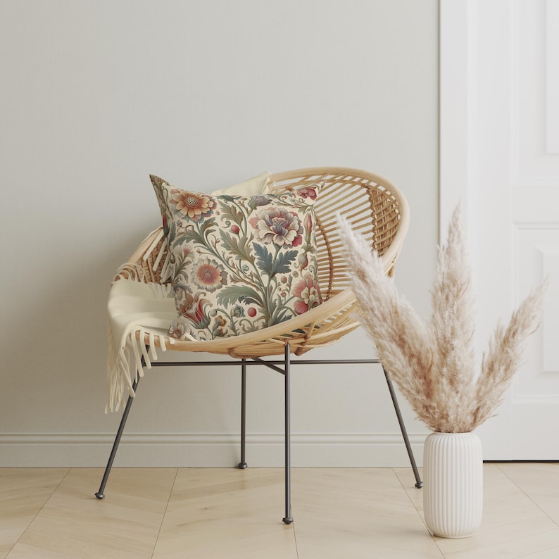 May include: A square throw pillow featuring a detailed botanical pattern with muted red, orange, and green floral motifs on a cream background, resting on a round rattan chair with thin black metal legs and a draped off-white throw blanket.