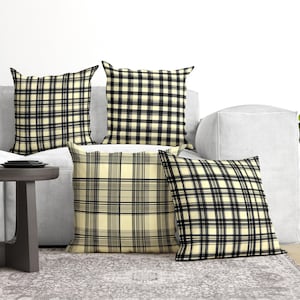 May include: Four square throw pillows featuring a plaid pattern in shades of cream, black, and grey. The pillows are arranged in a group, each displaying a consistent grid-like design with intersecting horizontal and vertical lines of varying thickness.