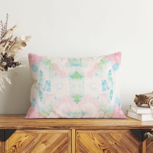 a wooden cabinet with a colorful, abstract pillow resting on top. The pillow has a watercolor-like design with shades of pink, blue, and green. The cabinet is situated against a white wall, and there is a vase of dried flowers on the left side of the frame.