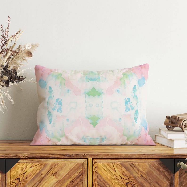 a wooden cabinet with a colorful, abstract pillow resting on top. The pillow has a watercolor-like design with shades of pink, blue, and green. The cabinet is situated against a white wall, and there is a vase of dried flowers on the left side of the frame.