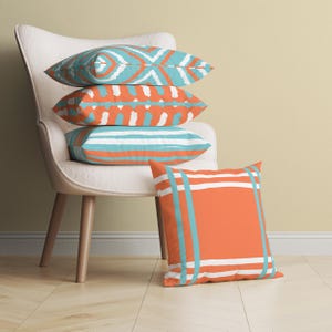 Puede incluir: Cuatro cojines decorativos con diseños en turquesa, naranja y blanco. Un cojín descansa en el suelo, mientras que los otros están apilados en una silla blanca con patas de madera. Los cojines presentan varios diseños geométricos y a rayas.