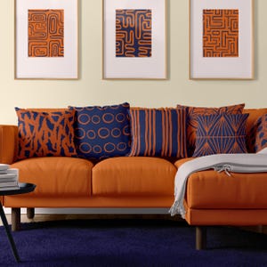 Op de afbeelding: Een oranje bank met vijf kussens met abstracte geometrische patronen in marineblauw. De kussens hebben verschillende ontwerpen, waaronder cirkels, strepen en zigzagpatronen. De bank staat voor een muur met drie ingelijste prints met vergelijkbare abstracte patronen.