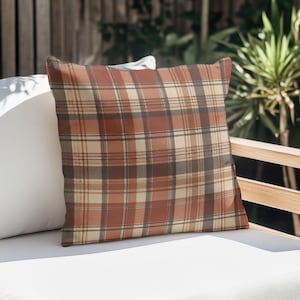 May include: A square throw pillow featuring a plaid pattern in shades of burnt orange, cream, and dark brown. The fabric displays a repeating grid of horizontal and vertical lines, creating a classic tartan design across the entire surface of the cushion.