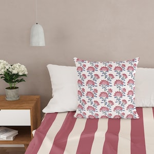 Puede incluir: Una almohada blanca con un estampado floral rosa y azul se encuentra sobre una cama con ropa de cama a rayas rosa y blanca.