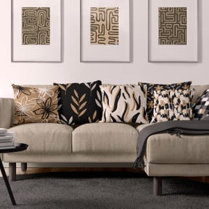 May include: A collection of five square throw cushions arranged on a beige sofa. The cushions feature various patterns in black, white, and tan, including floral motifs, leaf designs, abstract brushstrokes, and a spotted animal-inspired print.