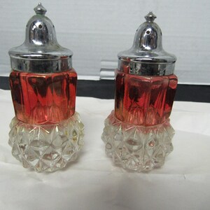 May include: A pair of vintage salt and pepper shakers. The shakers have a clear, faceted base, a red-tinted, geometric midsection, and a silver-colored metal top with a small, pointed finial. The shakers are displayed on a white surface.