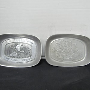 May include: Two silver-colored, rectangular trays. One tray has an embossed design with the words "Give Us This Day Our Daily Bread" and a pastoral scene. The other tray features a floral design.