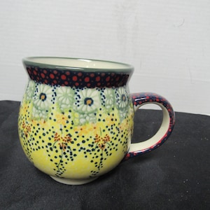 May include: A ceramic mug with a rounded shape and a handle. The mug features a yellow base adorned with small black dots and floral patterns in green, white, and yellow. The rim is dark blue with red dots, and the handle has a red and blue pattern.