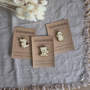 Könnte beinhalten: Drei Holz-Anstecknadeln auf Karten mit dem Text "TASCHENUMARMUNG" und "FÜHL DICH GEDRÜCKT". Die Anstecknadeln zeigen ein Krokodil, einen Bären und einen Pinguin. Die Karten tragen auch den Text "DIESE UMARMUNG IST FÜR DICH DA, WANN IMMER DU SIE BRAUCHST!"