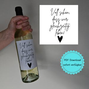 以下が含まれることがあります： 「Voll schön, dass wir gleichzeitig leben!」と書かれた白黒のラベルが貼られた白ワインのボトル。ラベルには小さな黒いハートも描かれている。