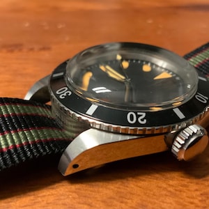Puede incluir: Un reloj de pulsera de acero inoxidable con esfera negra y bisel giratorio marcado con números blancos. El reloj tiene un cristal transparente y una correa de nailon a rayas verdes, rojas y negras. El reloj está sobre una superficie de madera.