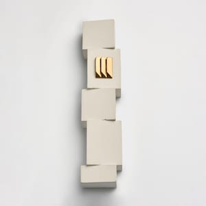 May include: A modern, abstract sculpture composed of stacked, off-white square blocks. A gold-colored, geometric accent is centered on one block. The sculpture is set against a plain white background, emphasizing its minimalist design.