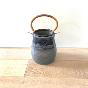 May include: A handcrafted ceramic vase with a blue and gray glaze. The vase features a woven handle and a decorative leaf design. The vase is approximately 7 inches tall and 6 inches wide.