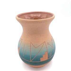 May include: A peach-coloured ceramic vase with a flared rim and a rounded body. The vase features a turquoise and teal gradient design with geometric shapes and arrows. The design is etched into the ceramic, creating a textured effect.