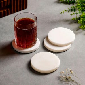 May include: A set of four circular white stone coasters with a smooth, matte finish. One coaster supports a clear glass tumbler filled with dark amber liquid, while the other three coasters are stacked or placed separately on a flat surface.