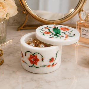 May include: A round white marble jewelry box with a removable lid. The exterior and lid feature inlaid stone designs of orange flowers, green vines, and a green bird. The open box contains gold rings with white pearls and clear stones.