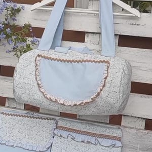 Puede incluir: Un bolso de bebé y accesorios a juego con un suave estampado de encaje floral. El bolso tiene un bolsillo y asas de color azul claro, con ribete marrón. Se incluyen artículos adicionales.