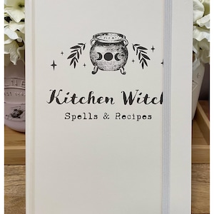 May include: Cream-colored notebook with the text "Kitchen Witch Spells & Recipes" and a cauldron illustration. The notebook has a gray elastic band closure and is surrounded by white flowers.