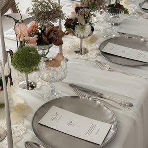 Könnte beinhalten: Elegante Tischdekoration mit weißer Tischdecke, silbernen Tellern, Besteck und Kristallgläsern. Blumengestecke in silbernen Vasen und hohe graue Kerzen tragen zum raffinierten Ambiente bei. Jedes Gedeck enthält eine Menükarte.