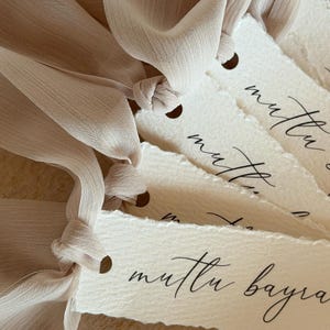 Puede incluir: Primer plano de varias etiquetas de papel blanquecino con un borde rasgado, cada una atada con una cinta beige claro. Las etiquetas tienen un peque&ntilde;o agujero en la parte superior y presentan una elegante escritura negra. Las etiquetas son probablemente para regalos u ocasiones especiales.