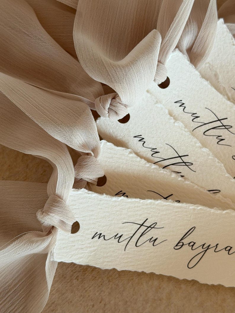 Puede incluir: Primer plano de varias etiquetas de papel blanquecino con un borde rasgado, cada una atada con una cinta beige claro. Las etiquetas tienen un peque&ntilde;o agujero en la parte superior y presentan una elegante escritura negra. Las etiquetas son probablemente para regalos u ocasiones especiales.
