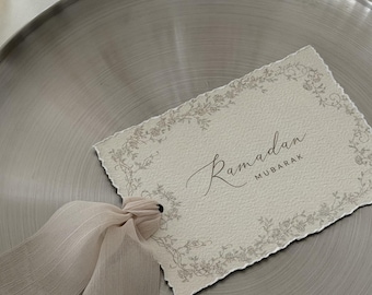 May include: A cream-coloured Ramadan Mubarak card with a floral border and a light pink ribbon, resting on a silver plate. The card's text is in elegant script.