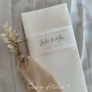 Puede incluir: Servilleta blanca con una etiqueta blanca que dice "Schon, dass du da bist Julia & Max Guten Appetit" y una cinta rosa atada alrededor. El texto "Sueño Atelier" está escrito en la servilleta.