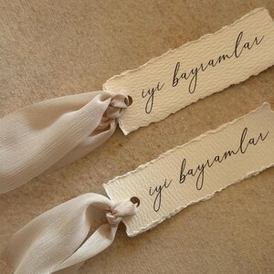 Puede incluir: Dos etiquetas de papel color crema con el texto manuscrito "iyi bayramlar" en escritura negra. Cada etiqueta tiene un peque&ntilde;o agujero y est&aacute; atada con una cinta color crema. El fondo es beige neutro.
