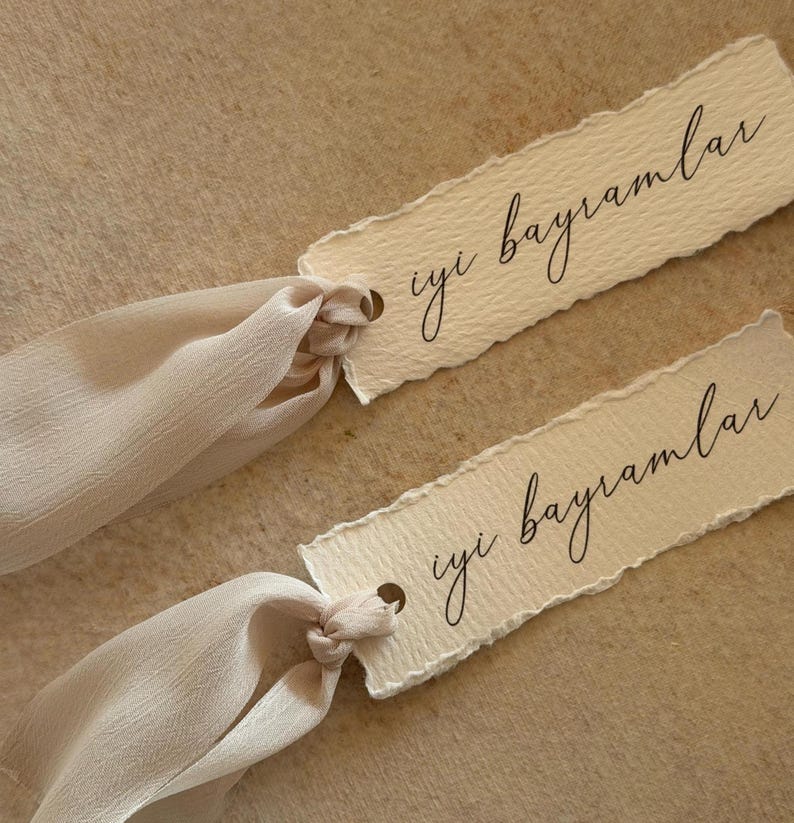Puede incluir: Dos etiquetas de papel color crema con el texto manuscrito "iyi bayramlar" en escritura negra. Cada etiqueta tiene un peque&ntilde;o agujero y est&aacute; atada con una cinta color crema. El fondo es beige neutro.