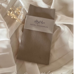 Könnte beinhalten: Ein gefaltetes, taupefarbenes Stoffstück, möglicherweise eine Serviette oder ein Tuch, ist mit einem hellgrauen Band umwickelt. Das Band zeigt eine elegante Schrift und die Worte "HAYIRLI RAMAZANLAR". Der Hintergrund ist ein weicher, weißer Stoff mit getrockneten floralen Akzenten.
