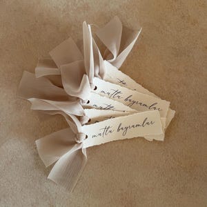 Puede incluir: Un mont&oacute;n de etiquetas de papel color crema con la frase "mutlu bayramlar" escrita en escritura negra. Cada etiqueta est&aacute; atada con una delicada cinta beige claro. Las etiquetas tienen un borde texturizado y rasgado.