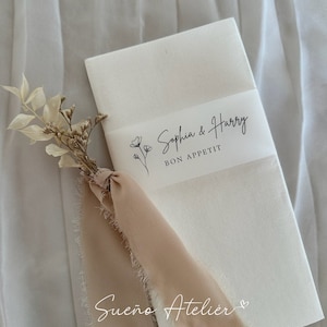 Puede incluir: Servilleta de papel blanca con una cinta blanca atada alrededor. La cinta tiene texto negro que dice "Sophia & Harry Buen provecho". Una pequeña rama de flores secas está unida a la cinta con una cinta beige.