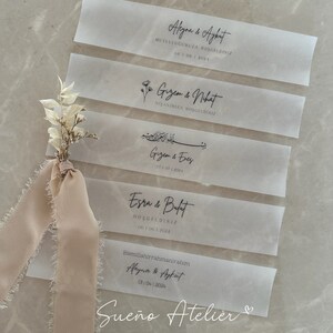 May include: Five white translucent paper tags with black calligraphy. The tags feature the names of couples and their wedding or engagement dates. The text on the tags reads: "Aleyna & Aykut", "Gizem & Nhat", "Gizem & Enes", "Esra & Bulat", and "Aleyna & Ayfut". The bottom tag also includes the text "Sue&ntilde;o Atelier" and a heart symbol.