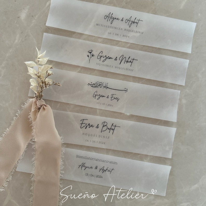 May include: Five white translucent paper tags with black calligraphy. The tags feature the names of couples and their wedding or engagement dates. The text on the tags reads: "Aleyna & Aykut", "Gizem & Nhat", "Gizem & Enes", "Esra & Bulat", and "Aleyna & Ayfut". The bottom tag also includes the text "Sue&ntilde;o Atelier" and a heart symbol.