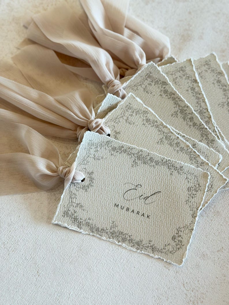 Pu&ograve; includere: Un set di etichette rettangolari in carta con bordi sfrangiati, ciascuna caratterizzata da un bordo floreale grigio e dal testo Eid MUBARAK al centro. Ogni etichetta &egrave; fissata a un lungo e fluido nastro di chiffon beige annodato.