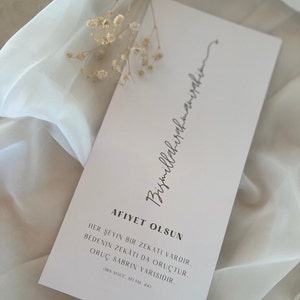 May include: A rectangular card with text in a script font, with the words "Bismillahirahmanirahim" at the top. The card also has the words "AFIYET OLSUN" and other text in a script font. The card is light pink and is decorated with dried flowers.